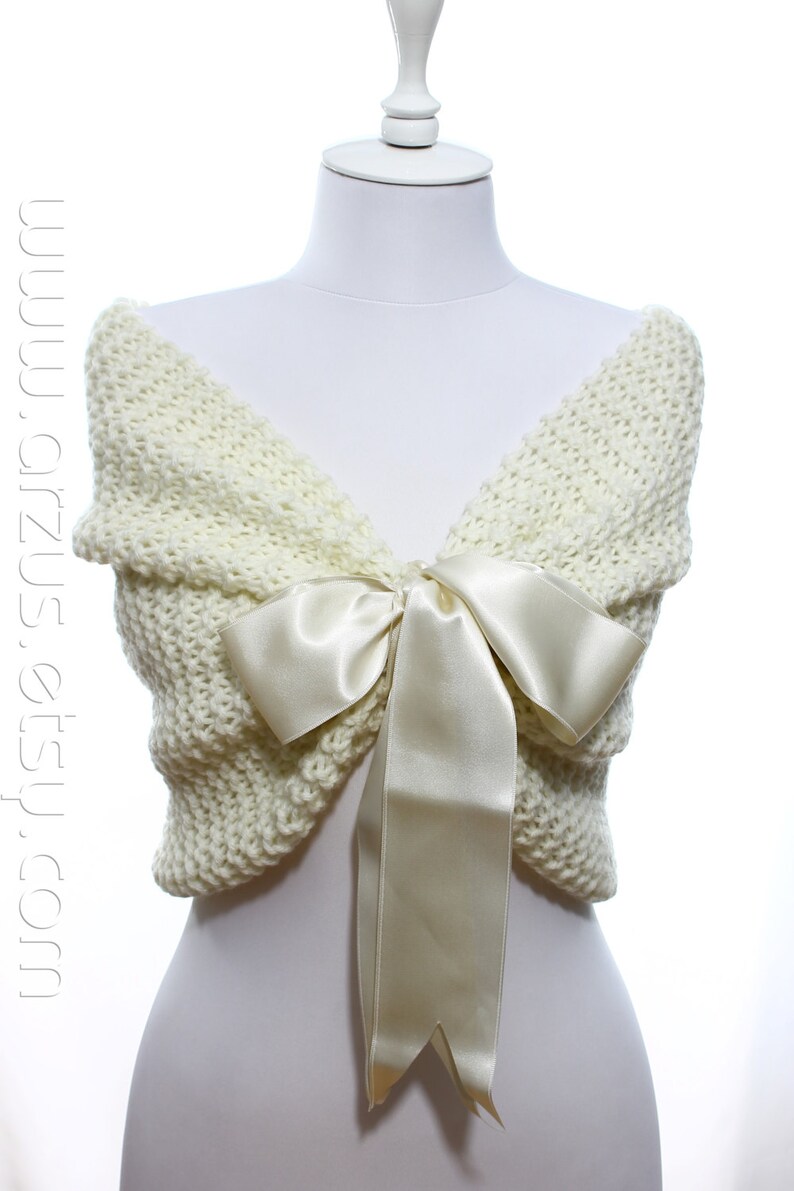 May include: A white knitted shawl with a large ivory satin bow. The shawl is draped over a white mannequin.