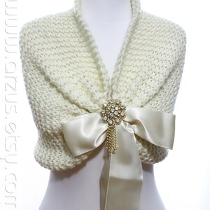 May include: A white knitted shawl with a satin bow and a rhinestone brooch. The shawl is draped over a white mannequin.