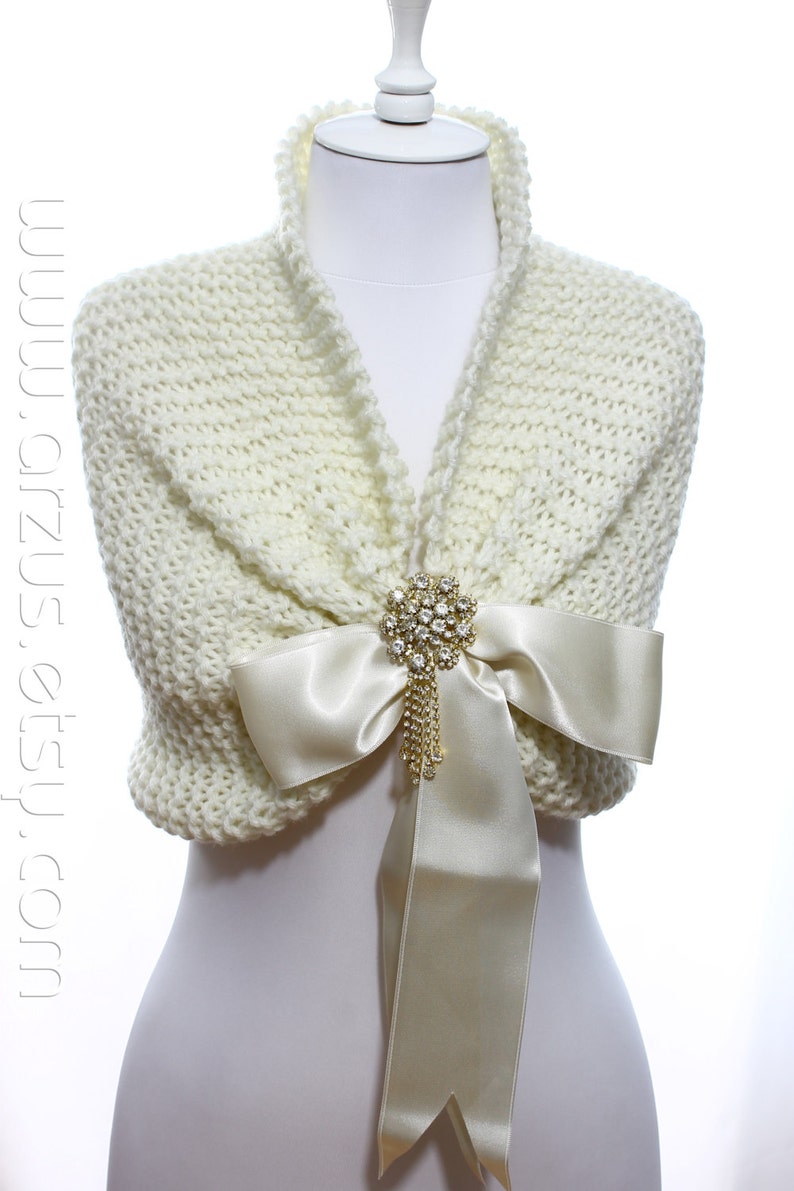 May include: A white knitted shawl with a satin bow and a rhinestone brooch. The shawl is draped over a white mannequin.