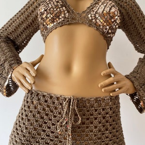 May include: A brown crochet top with long sleeves and a brown crochet skirt. The top has gold sequins and a cutout detail at the neckline. The skirt has a tie at the waist.