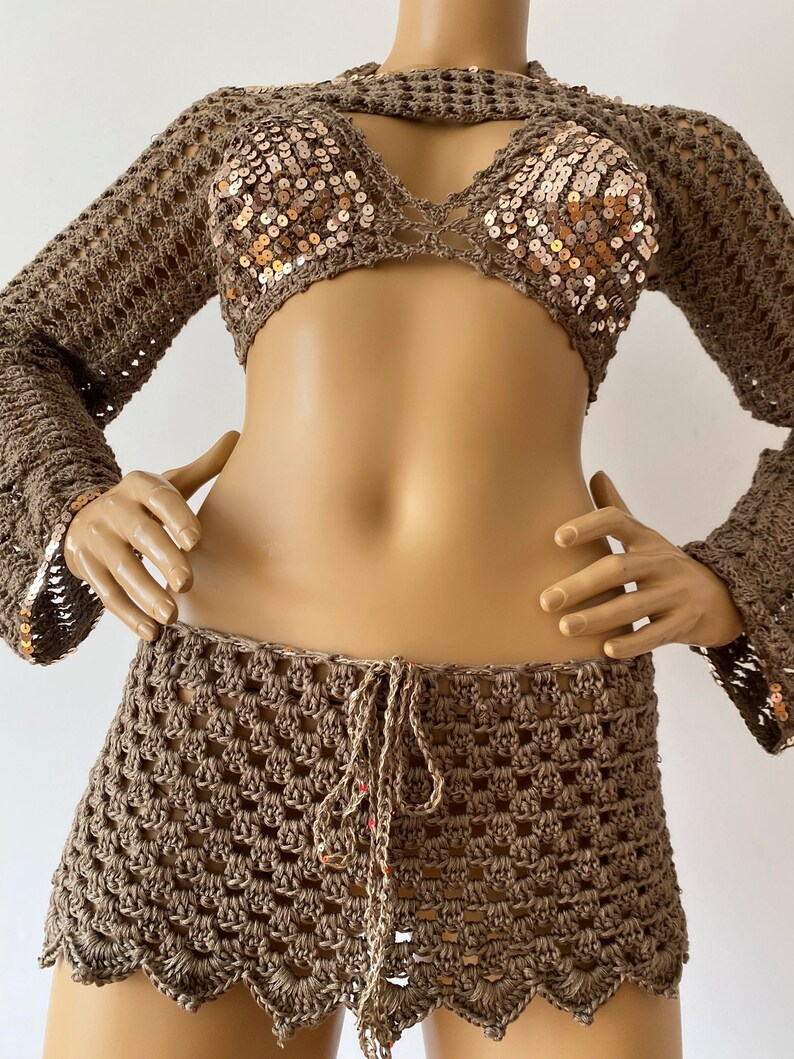 May include: A brown crochet top with long sleeves and a brown crochet skirt. The top has gold sequins and a cutout detail at the neckline. The skirt has a tie at the waist.