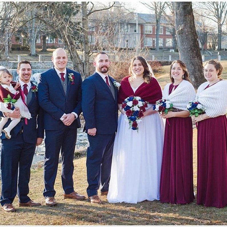 May include: A wedding party poses for a photo in front of a building. The bride is wearing a white dress with a red shawl and a bouquet of flowers. The groom is wearing a dark blue suit and a white shirt. The bridesmaids are wearing burgundy dresses and white shawls. The groomsmen are wearing dark blue suits and white shirts. A young girl is holding a bouquet of flowers and wearing a white dress.