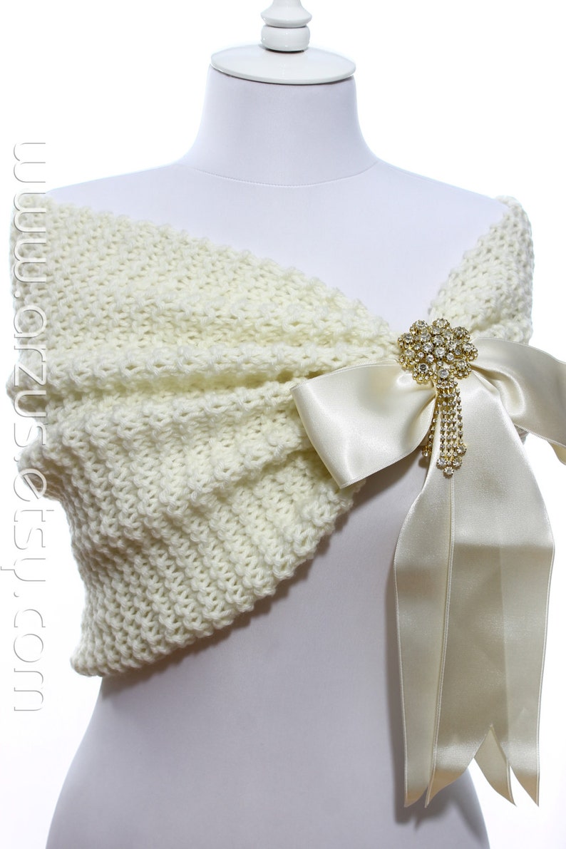 May include: A white knitted shawl with a large ivory satin bow and a rhinestone brooch. The shawl is draped over a white mannequin.