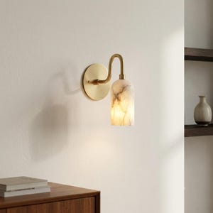 May include: A wall-mounted light fixture featuring a circular brass backplate and a curved arm that supports a cylindrical, translucent stone shade with natural grey veining, emitting a warm, soft glow from within the stone structure.
