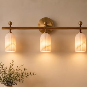 May include: A three-light wall fixture featuring a horizontal brass bar with a circular mounting plate. Three cylindrical, vertically ribbed white glass shades are suspended from the bar, each topped with a small brass sphere finial.