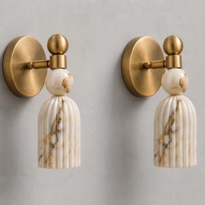 May include: A pair of wall sconces featuring a circular brushed brass backplate, a short horizontal arm, and a vertical fixture composed of a smooth spherical accent atop a fluted, cylindrical white marble shade with brown veining.