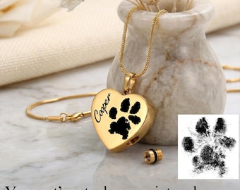May include: A gold-colored heart-shaped pendant necklace with a black paw print and the name "Cooper" in script. The necklace includes a small screw-off compartment and a matching gold chain. The image also shows a paw print and the text "Your pet's actual paw-print and name."