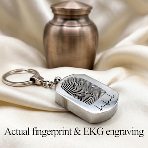 May include: A silver keychain with a fingerprint and EKG engraving, next to a bronze urn. The keychain has a rectangular tag with a detailed fingerprint design and a small EKG line. The urn is a rounded shape with a lid.