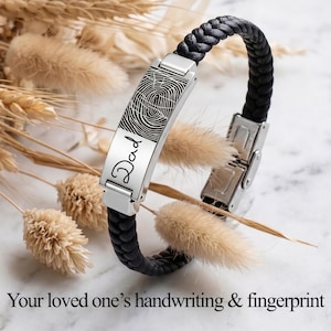 May include: A black braided leather bracelet featuring a rectangular silver-tone metal plate engraved with a black fingerprint pattern and the word Dad in cursive script. The bracelet includes a silver-tone metal clasp mechanism.