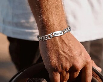 Puede incluir: Una pulsera de cadena plateada con una placa rectangular grabada con el nombre "David". La pulsera se lleva en la muñeca, con la mano sosteniendo un objeto. La pulsera tiene un diseño de eslabones gruesos.