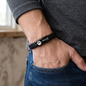 May include: A black braided leather bracelet featuring a rectangular metal clasp. The clasp displays a white engraved paw print icon on one side and the name Mocca in a white script font on the adjacent side of the black metal surface.
