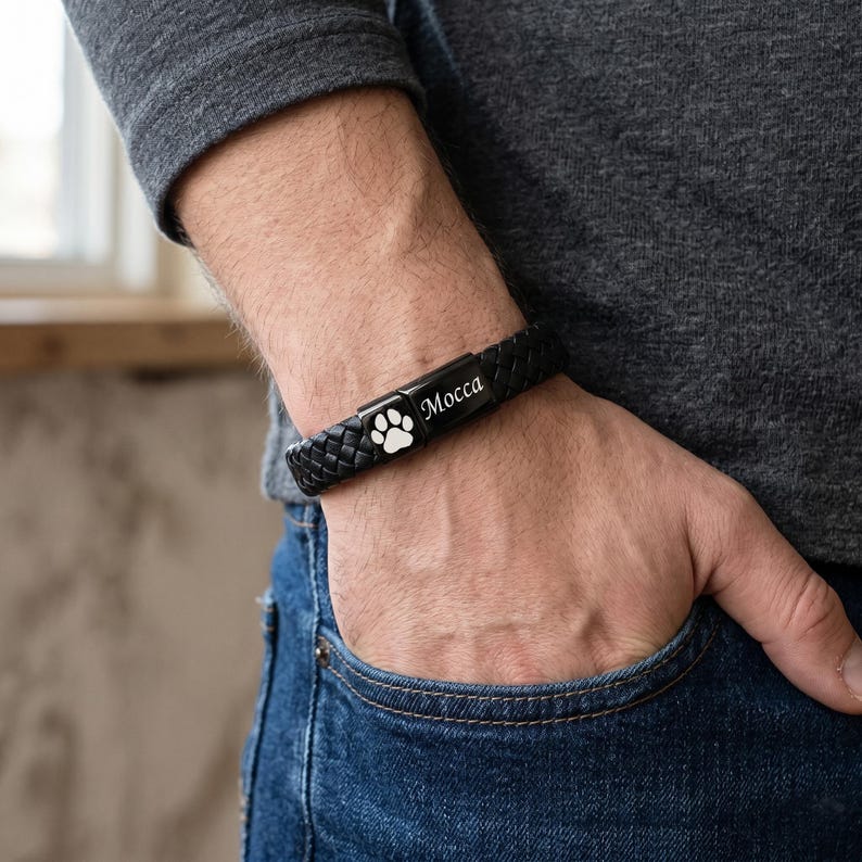 May include: A black braided leather bracelet featuring a rectangular metal clasp. The clasp displays a white engraved paw print icon on one side and the name Mocca in a white script font on the adjacent side of the black metal surface.