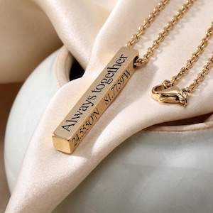 May include: A gold-colored necklace with a rectangular pendant. The pendant is engraved with the words "Always together" and coordinates 24.5550°N 81.7759°W. The necklace is displayed on a cream-colored fabric.