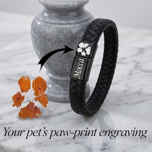 May include: A black braided leather bracelet featuring a rectangular black metal clasp. The clasp is engraved with a white paw print and the name Mocca in a script font. The text Your pet's paw-print engraving is displayed below the bracelet.