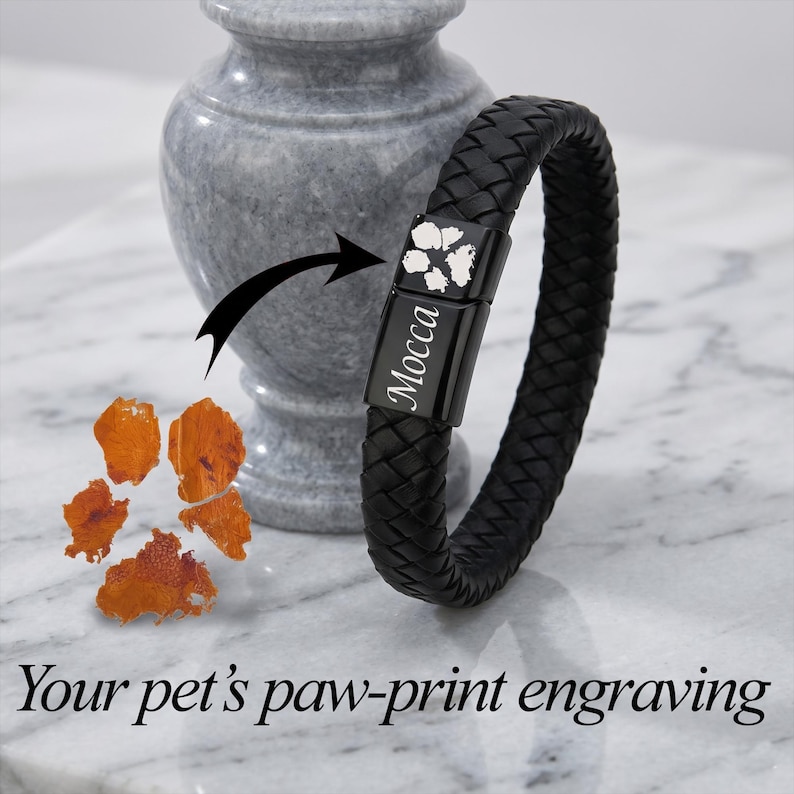 May include: A black braided leather bracelet featuring a rectangular black metal clasp. The clasp is engraved with a white paw print and the name Mocca in a script font. The text Your pet's paw-print engraving is displayed below the bracelet.