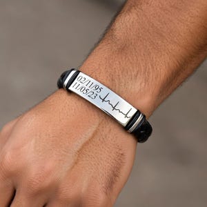 May include: A black leather bracelet with a silver plate. The plate has the dates 02/11/95 and 11/05/23, along with a heartbeat line design. The bracelet is a stylish accessory.