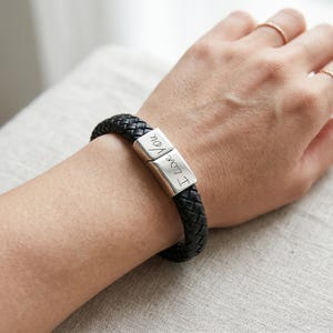 May include: A black braided leather bracelet featuring a rectangular silver-toned metal clasp. The clasp is engraved with the cursive text I Love You, split across two segments that connect to secure the bracelet around the wrist.