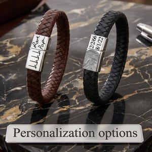 May include: Two braided leather bracelets, one brown and one black, each featuring a rectangular silver-toned metal clasp. The brown bracelet clasp displays a handwritten signature and heartbeat line. The black bracelet clasp shows a fingerprint and two dates.