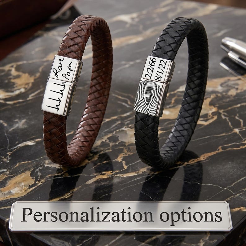 May include: Two braided leather bracelets, one brown and one black, each featuring a rectangular silver-toned metal clasp. The brown bracelet clasp displays a handwritten signature and heartbeat line. The black bracelet clasp shows a fingerprint and two dates.