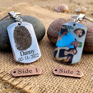 May include: Two personalized dog tag keychains. One tag features a fingerprint and the name "Danny" with a date. The other tag displays a color photograph of three people. Both tags have a silver keyring and are on a textured surface.