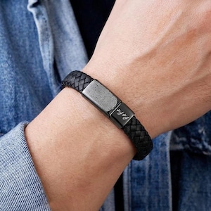 Può includere: Braccialetto in pelle intrecciata nera con un design a impronta digitale su una chiusura rettangolare in metallo. La chiusura presenta anche il testo inciso "P&Fe". Il braccialetto è indossato al polso.