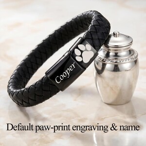 May include: A black braided leather bracelet with a rectangular black metal clasp featuring an engraved paw print and the name Cooper, positioned next to a small, polished silver-toned metal urn with a lid and a decorative rope-textured rim.