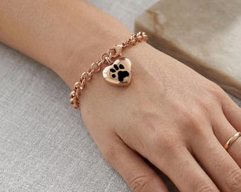 May include: Rose gold bracelet featuring a heart-shaped charm with a black paw print. The charm is suspended from a chain-link bracelet. The bracelet is worn on a wrist, with a simple gold ring on a finger.