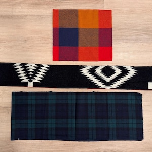May include: Three rectangular fabric swatches: one with a multi-colored orange, red, and blue plaid pattern, one long black strip featuring a white geometric diamond and tree design, and one with a dark blue and green tartan plaid pattern.