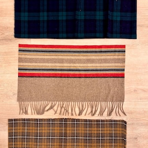 May include: Three rectangular scarves arranged vertically. The top scarf features a dark blue and green plaid pattern. The middle scarf has horizontal stripes in red, gray, and tan with a fringed edge. The bottom scarf has a brown plaid pattern.