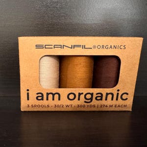 May include: A rectangular cardboard box containing three spools of thread in cream, mustard yellow, and dark brown. The packaging is labeled SCANFIL ORGANICS and i am organic, with text indicating 3 spools, 30/2 WT, and 300 YDS per spool.
