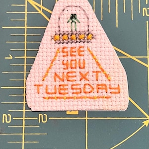 May include: A pink triangle-shaped fabric patch with orange embroidery that reads "See you next Tuesday". A green stick figure is embroidered in the top of the triangle.