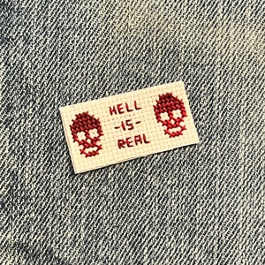 May include: A white cross-stitch patch with red embroidered skulls and the text "HELL IS REAL".