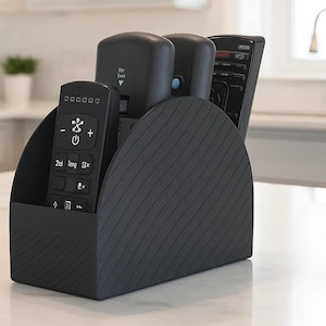 May include: A black plastic remote control organizer with a curved side profile and diagonal textured lines. The holder contains three black remote controls of varying heights, with the front-most remote displaying white button icons and text.