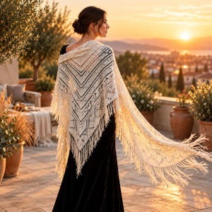 May include: A cream-colored triangular shawl featuring an intricate openwork knit pattern and long fringe along the edges. The shawl is draped over the shoulders, showcasing a detailed geometric design with delicate lace-like holes.