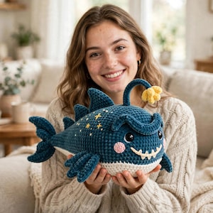 Puede incluir: Un peluche de rape tejido a ganchillo en color azul oscuro con vientre crema, un señuelo amarillo, detalles de estrellas bordadas en el lomo, mejillas rosadas y una amplia sonrisa con dientes blancos visibles, hecho de hilo suave y texturizado.
