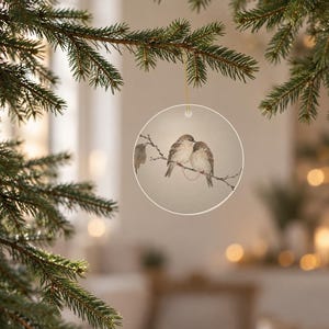May include: A circular white ceramic ornament featuring a detailed illustration of two small brown birds perched together on a thin branch. A delicate red line connects the two birds, and the ornament hangs from a thin gold string.