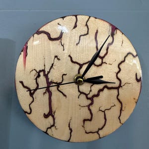 May include: A circular wall clock featuring a light-colored wood face with dark, branching fractal patterns and purple accents. The clock has three thin black hands centered on a gold-toned mechanism, creating a high-contrast, organic design.