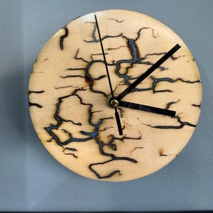 May include: A circular wall clock featuring a light-colored wooden face with dark, intricate fractal burn patterns resembling lightning or tree branches. The clock has three thin black hands centered on the face, with no numbers or markings.
