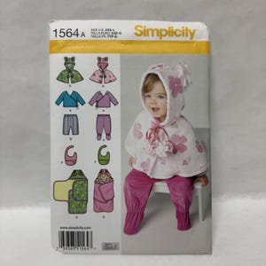 May include: Simplicity sewing pattern 1564A for infant clothing, including a hooded cape with floral details, pants, and a swaddle wrap. The envelope displays illustrations of various garment options in sizes XXS-L, featuring pink and green color schemes.