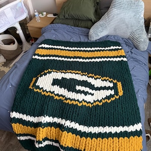 May include: A chunky knit blanket featuring a large white letter G logo centred on a dark green background. The blanket has horizontal stripes of yellow and white at the top and bottom edges, crafted from thick, soft yarn in a textured pattern.