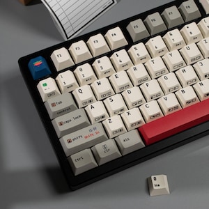 Puede incluir: Un teclado mecánico con marco negro y teclas color hueso con letras y símbolos negros. Incluye una tecla escape azul oscuro con un gráfico, una barra espaciadora roja y teclas modificadoras grises. Una tecla 0 ins suelta se encuentra cerca.