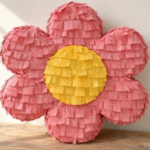 Pode incluir: Uma piñata em forma de flor com um centro circular amarelo brilhante rodeado por seis pétalas cor-de-rosa arredondadas. Toda a superfície é coberta por camadas de papel crepe com franjas, criando uma aparência texturizada e em camadas.