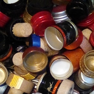 May include: A collection of various bottle closures, including corks, metal screw caps, and plastic caps. The caps are in a variety of colors, including red, black, green, and gold. Some caps have labels or designs. The image is a close-up shot.