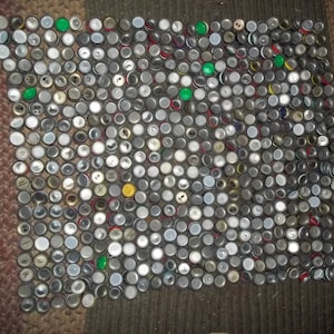May include: A large collection of bottle caps, mostly silver, with a few green and yellow ones, arranged in a grid pattern on a brown and white striped surface.