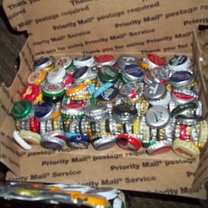 May include: A close-up image of a box filled with a large collection of bottle caps. The bottle caps are a variety of colors, including gold, silver, red, blue, and green. The caps are arranged in a random pattern, and some of them are stacked on top of each other.