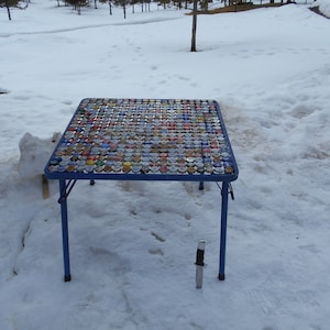 May include: A blue folding table with a top made of bottle caps. The table is sitting on a snowy surface.