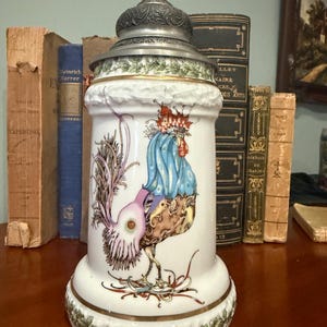 May include: A white ceramic beer stein featuring a colourful illustration of a rooster with blue, purple, and red plumage. The stein has a metallic pewter lid with an ornate finial and a green leaf pattern border at the base and top rim.