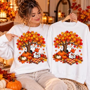 Puede incluir: Dos sudaderas blancas de cuello redondo con un estampado gráfico de un perro de dibujos animados sentado bajo un gran árbol de otoño con hojas naranjas y amarillas. El diseño incluye calabazas, una manta a cuadros y una taza roja.