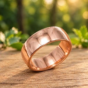 May include: A polished rose gold band ring with a smooth, reflective surface. The ring features a rounded exterior profile and a plain, unadorned design, showcasing the warm, metallic pinkish-copper hue of the metal in a simple, circular form.