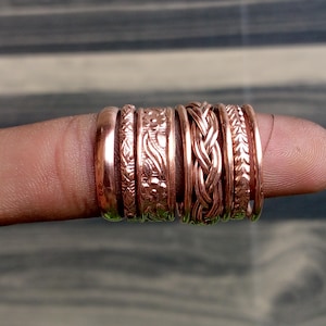 May include: A set of six copper rings featuring various textures and patterns, including a smooth band, a floral embossed band, a braided design, and a leaf-like pattern, all displayed together in a row on a finger.